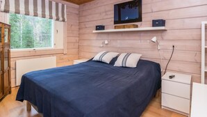 A bedroom with a wooden ceiling, a bed with a dark blue bedspread, a window with a striped valance, a nightstand, and a shelf above the bed. - Isokuru 3 by Interhome (Pelkosenniemi)
