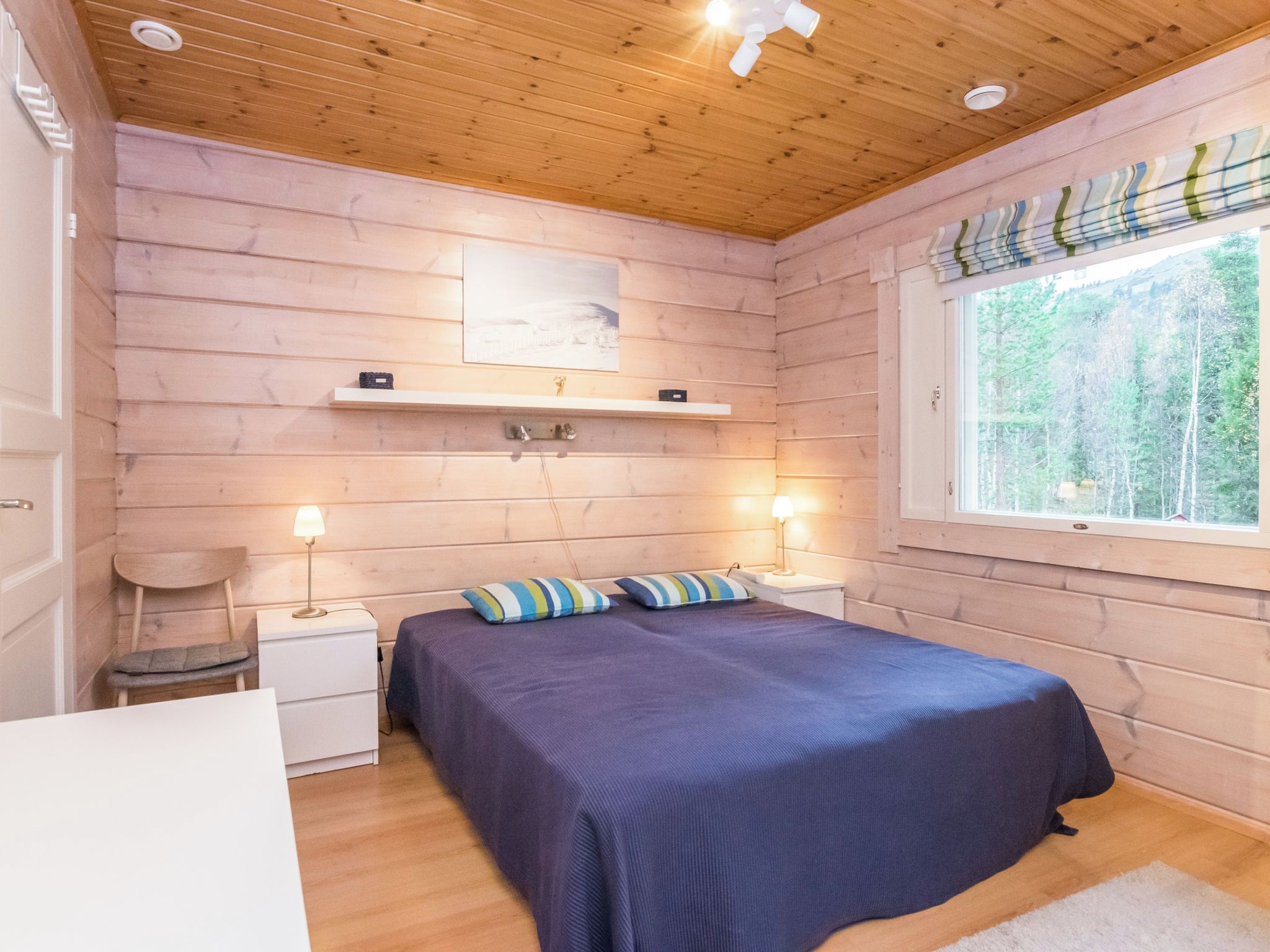 Une chambre avec un plafond en bois, un lit avec une literie bleue, deux tables de chevet avec des lampes, une fenêtre donnant sur des arbres et une étagère avec un tableau.