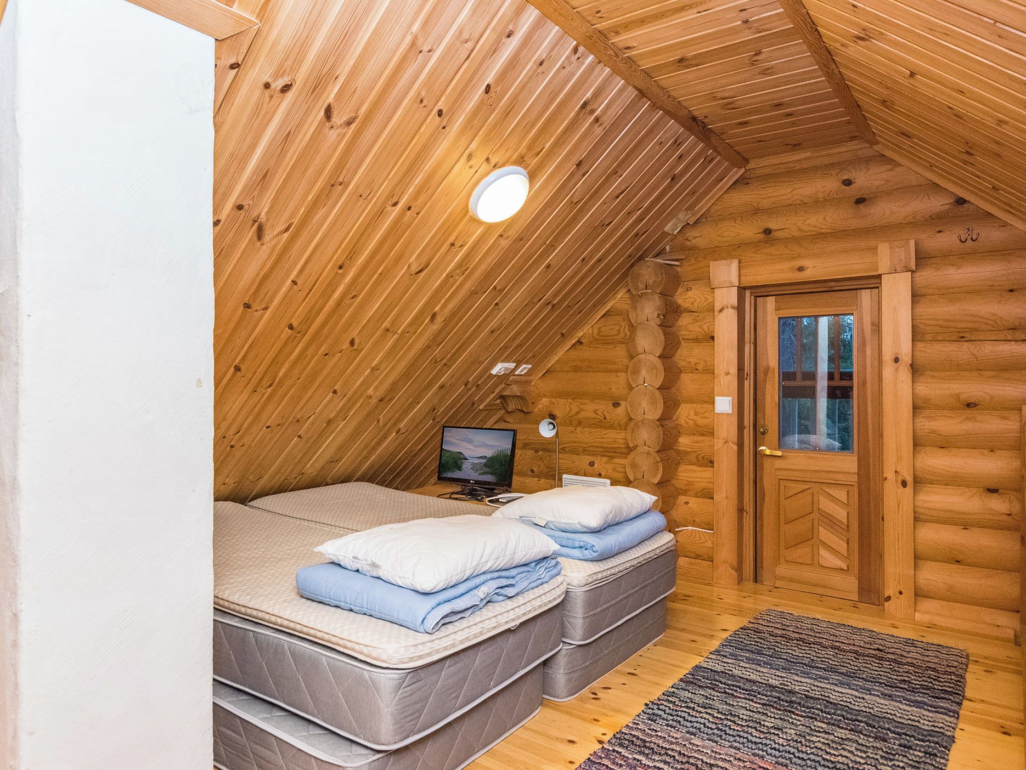 Un dormitorio tipo cabaña de madera con techo inclinado, un televisor sobre un mueble, dos camas con almohadas y una puerta de madera.