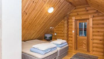 Un dormitorio tipo cabaña de madera con techo inclinado, un televisor sobre un mueble, dos camas con almohadas y una puerta de madera.