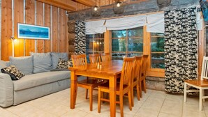 A cozy cabin interior with a wooden dining table, chairs, a sofa, and a large window with curtains.