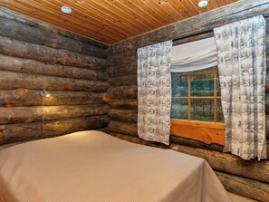 A log cabin room with a bed, a window with curtains, and a wall-mounted light.