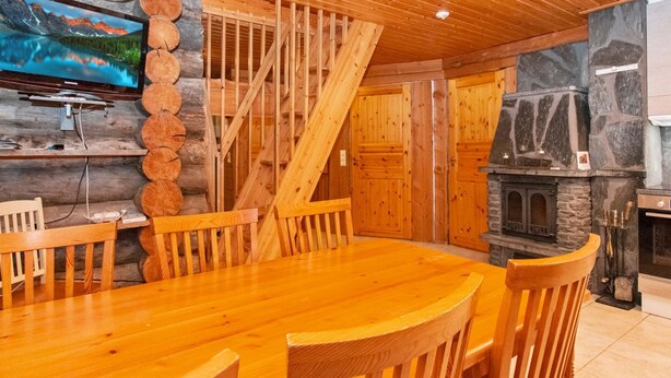 A wooden interior with a dining table, chairs, a fireplace, and a television mounted on a log wall.