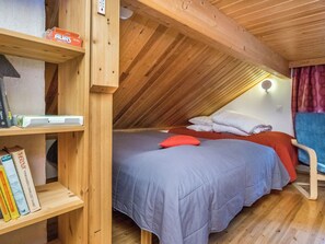 Une chambre mansardée chaleureuse avec un plafond en pente, un lit recouvert d’une couverture bleue et agrémenté d’un oreiller rouge, une étagère en bois remplie de livres et une petite table avec une lampe.