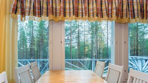 A wooden dining table with white chairs, a plaid curtain, and a view of a forest through large windows. - Isokurunpuisto a 2 by Interhome (Pelkosenniemi)