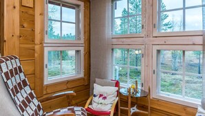 L’intérieur chaleureux d’un chalet avec des murs en bois, un fauteuil à carreaux, une petite table et de nombreuses fenêtres donnant sur une zone boisée.