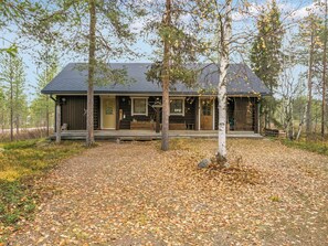 A log cabin with a porch, surrounded by trees and a leaf-covered ground.