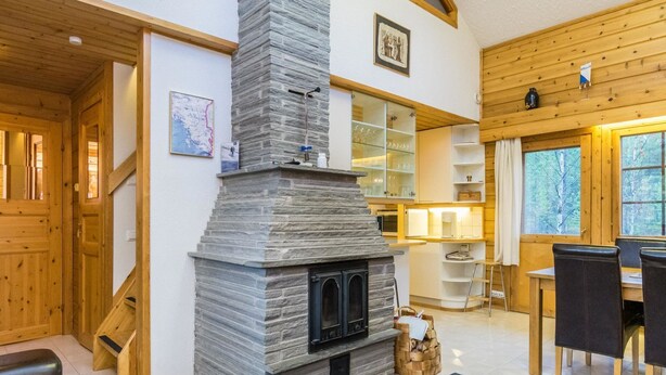 A cozy interior with a stone fireplace, wooden walls, a dining area with chairs, and a staircase.