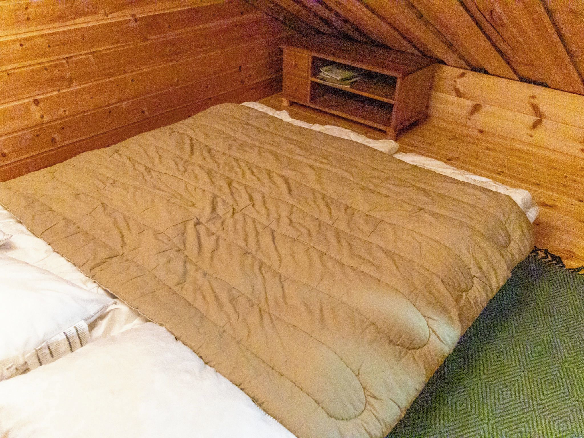 Een houten kamer met een bed, een klein houten kistje en een vloerkleed met een groen patroon.