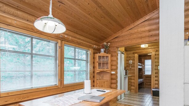 A wooden cabin interior with a large dining table, wooden benches, a kitchen area with a refrigerator, and a window with blinds.