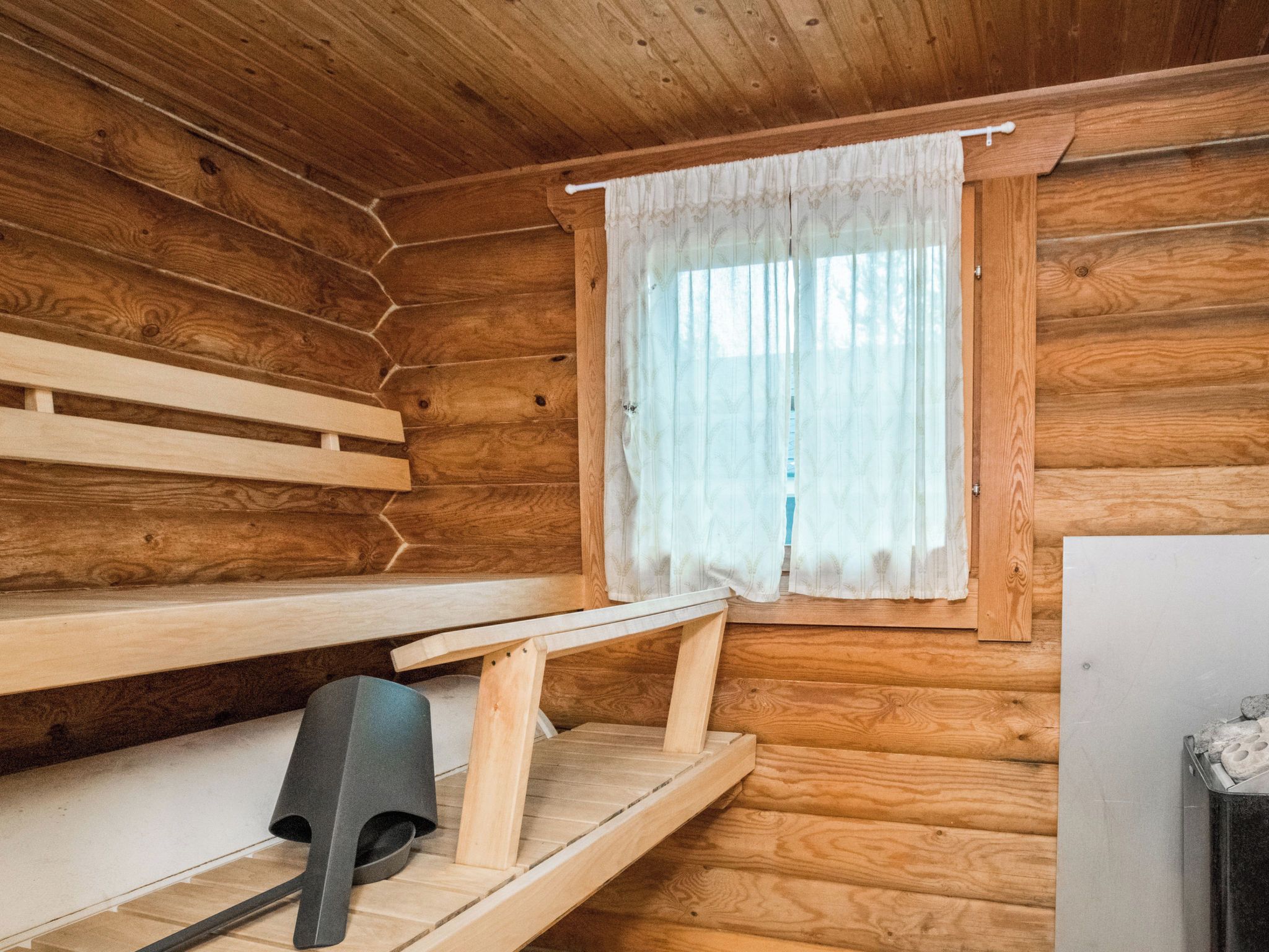 Interior de una cabaña de madera con un banco, una mesita y una ventana con cortinas.