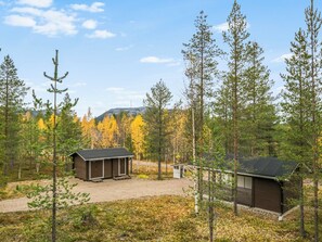 A cabin with a small porch and a separate shed, surrounded by a forest with autumn-colored trees.
