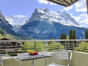 Een balkon met tafel en stoelen, met uitzicht op een bergachtig landschap met besneeuwde toppen en groene bomen.