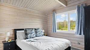 A bedroom with a wooden ceiling, a large window with curtains, a bed with white bedding, and a nightstand with a lamp and books. - Chestnut Lodge - Pet Friendly - Free Parking & WiFi (Exeter)