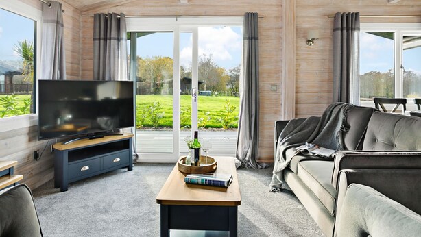 A modern living room with a flat-screen TV, a sofa, a coffee table, and large windows offering a view of a green garden. - Chestnut Lodge - Pet Friendly - Free Parking & WiFi (Exeter)