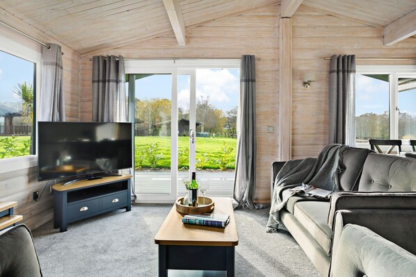 Ein modernes Wohnzimmer mit Flachbildfernseher, einer Couch, einem Couchtisch und großen Fenstern mit Blick auf einen grünen Garten.