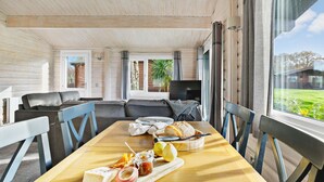 A cozy living room with a wooden ceiling, a grey sofa, a wooden dining table with food, and a TV in the background. - Chestnut Lodge - Pet Friendly - Free Parking & WiFi (Exeter)