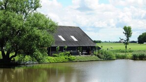 Ein rustikales Bauernhaus mit geneigtem Dach und zwei Dachfenstern, neben einem ruhigen Teich mit üppigem Grün und blauem Himmel.