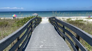 Ein hölzerner Steg führt zu einem Strand mit blauem Himmel und weiter Meerblick.
