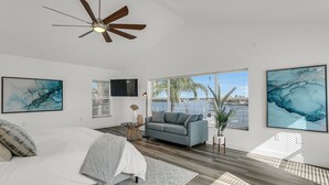 Ein Schlafzimmer mit einem Bett, einem grauen Sofa, einem Deckenventilator, einem Fernseher und großen Fenstern mit Blick auf das Wasser.