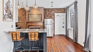 Une cuisine avec un mur en briques, des tabourets de bar en bois, un îlot central en acier inoxydable, des luminaires suspendus et un réfrigérateur blanc.