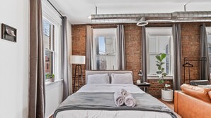 Une chambre d’hôtel moderne avec un grand lit, un canapé en cuir et une vue sur la ville grâce à de grandes fenêtres.