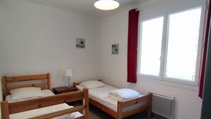 A room with two wooden beds, a small bedside table with a lamp, a radiator, and a window with red curtains. - Quiet house near Domino market (Saint-Georges-d'Oléron)