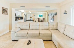 A living room with a beige sectional sofa, a coffee table with remotes, and a kitchen in the background.