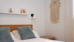 Un letto ben rifatto con lenzuola bianche e asciugamani piegati, una testiera in legno, un comodino con una lampada e una decorazione in macramè appesa al muro.