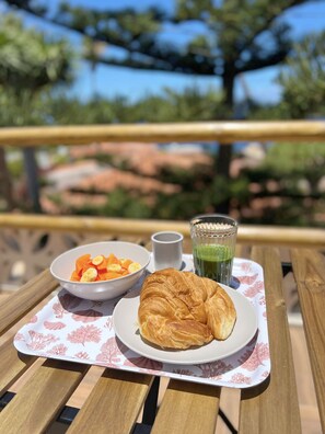A table with a croissant, a bowl of fruit, and a glass of green juice on a wooden table outdoors. - Apartment with a terrace overlooking the ocean and the sunset (Bajamar)