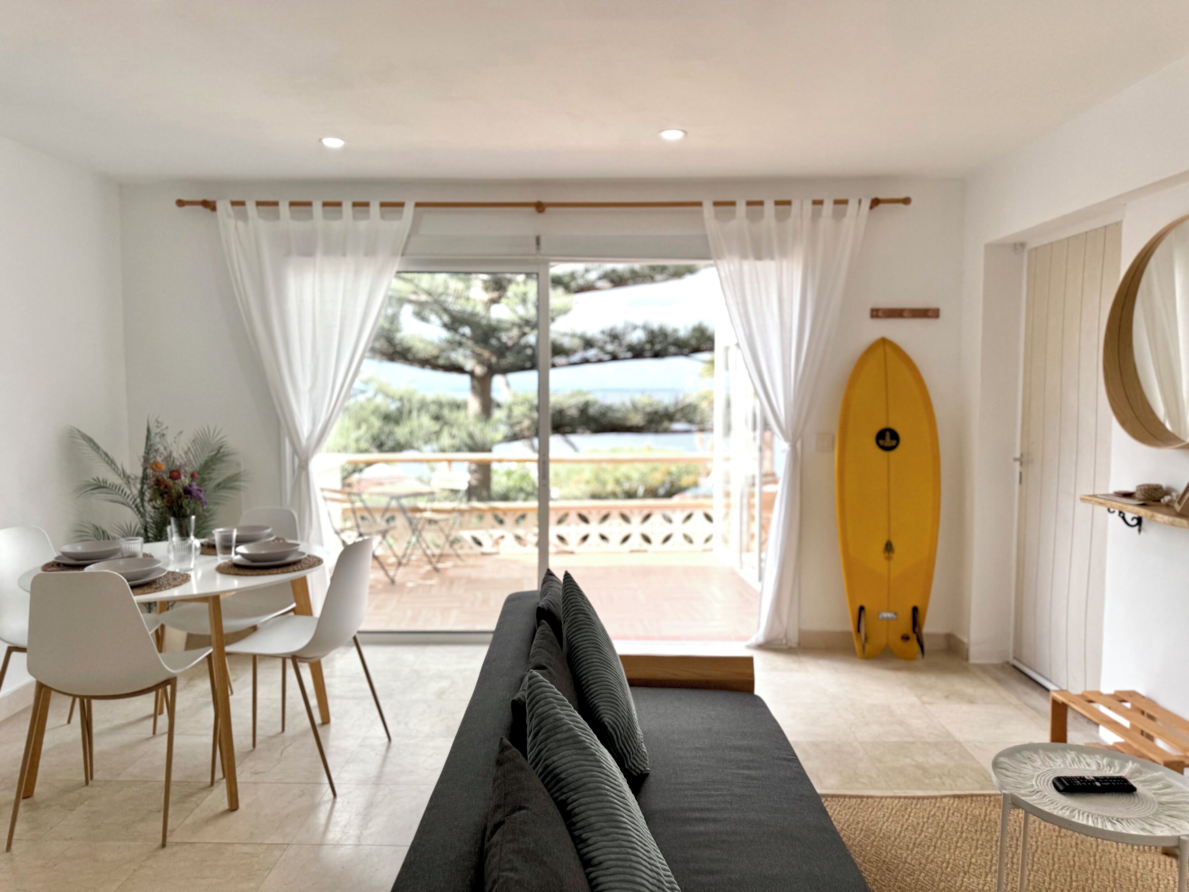 A modern living room with a sofa, dining table, chairs, and a surfboard.