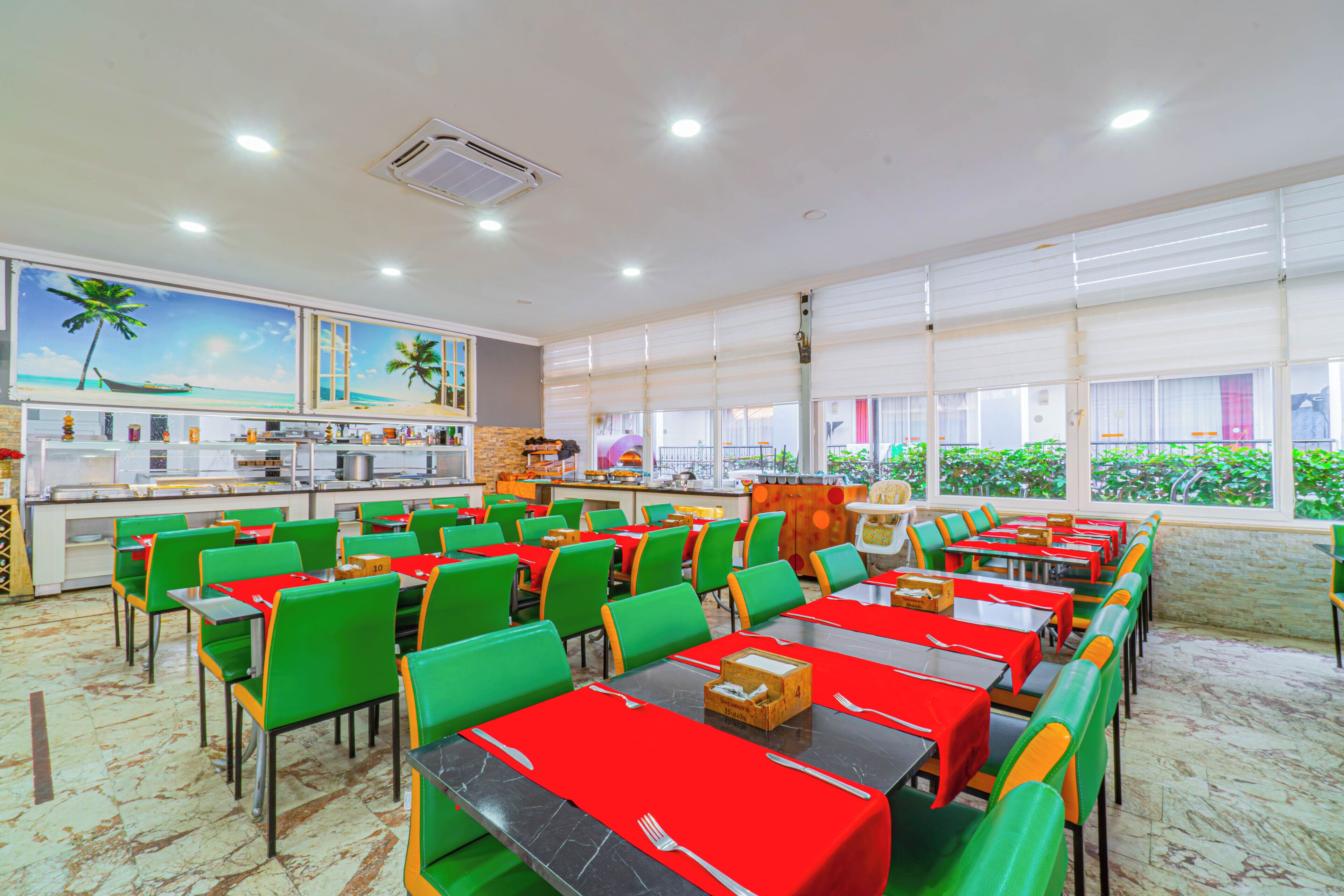 A spacious dining area with multiple tables and chairs, a kitchen in the background, and large windows with a view of greenery.