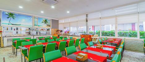 A spacious dining area with multiple tables and chairs, a kitchen in the background, and large windows with a view of greenery.