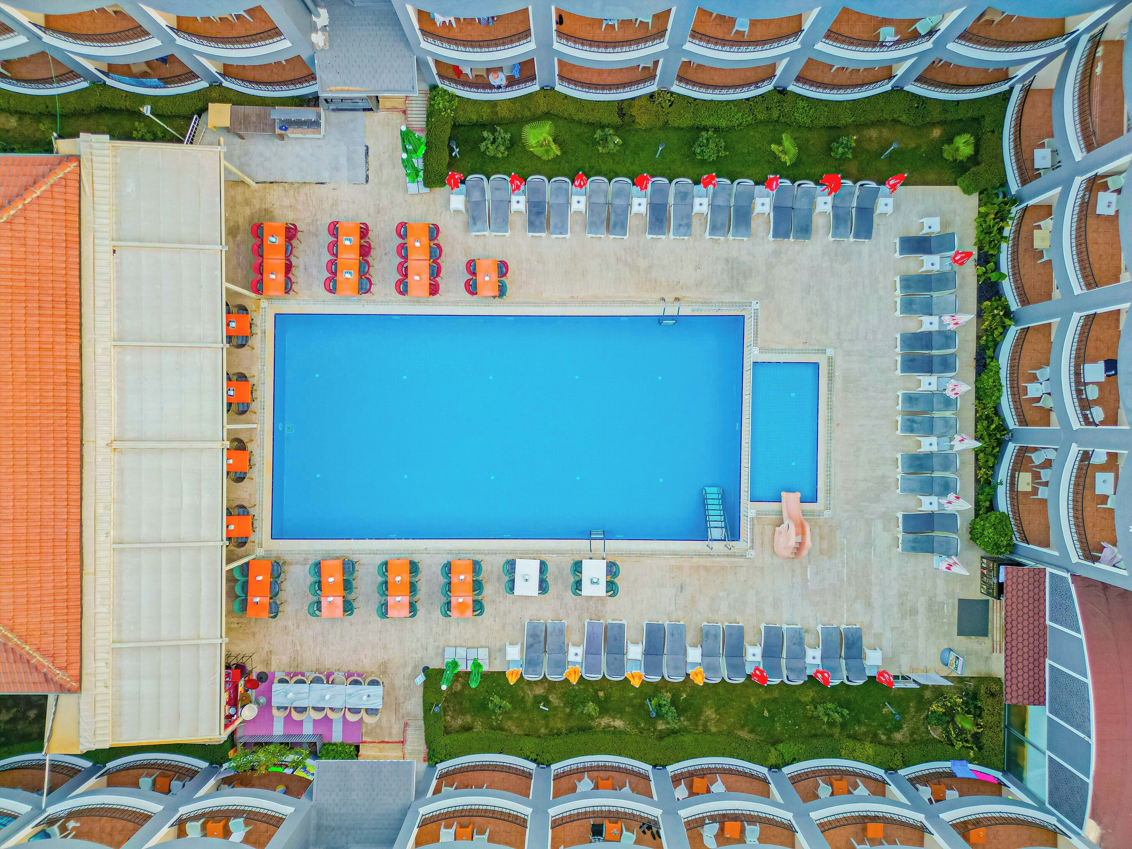 An aerial view of a hotel with a large rectangular pool, multiple lounge areas with chairs and umbrellas, and a central dining area.