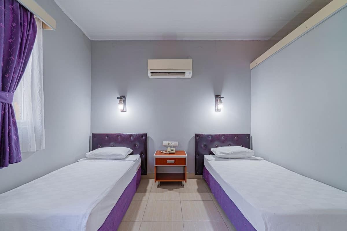 A hotel room with two single beds, a small wooden nightstand, a wall-mounted air conditioner, and two wall-mounted lights.