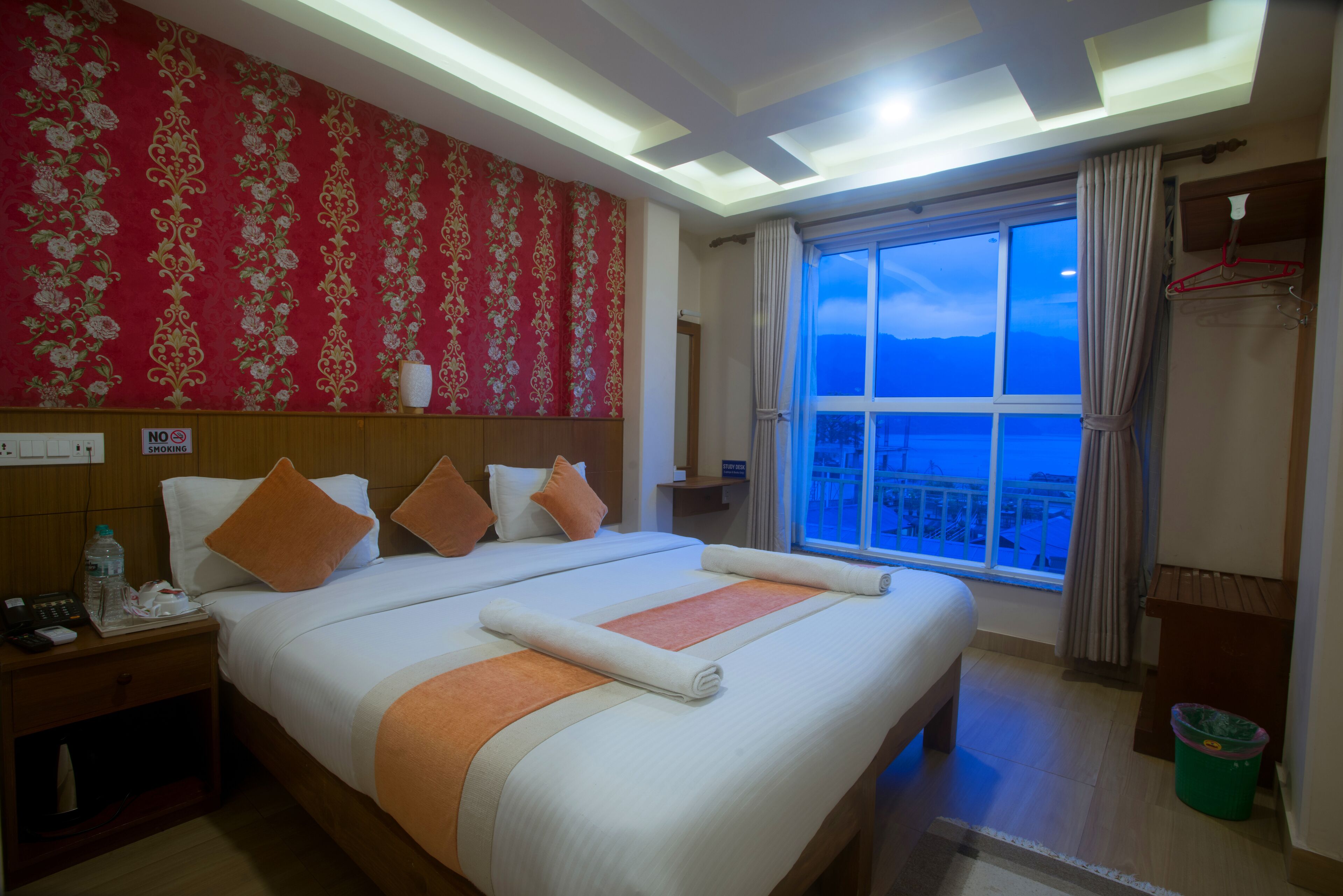 A hotel room with a large bed, a nightstand, a window with a view, and a red patterned wall.