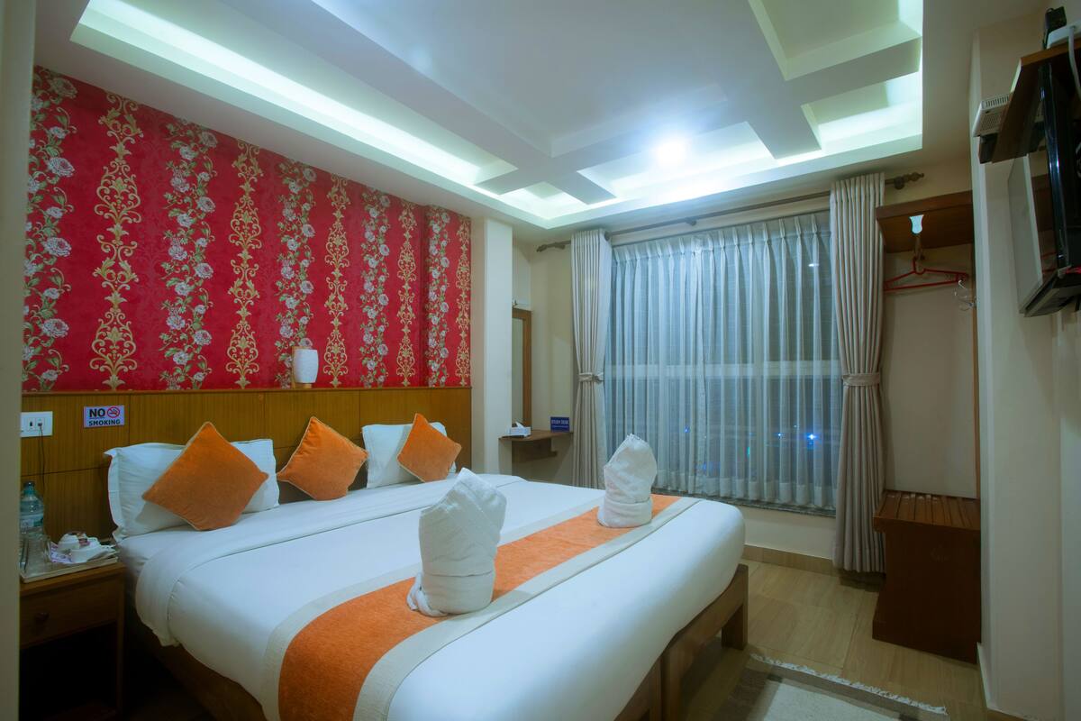 A hotel room with a large bed, a red floral wallpaper, a window with curtains, and a television on the wall.