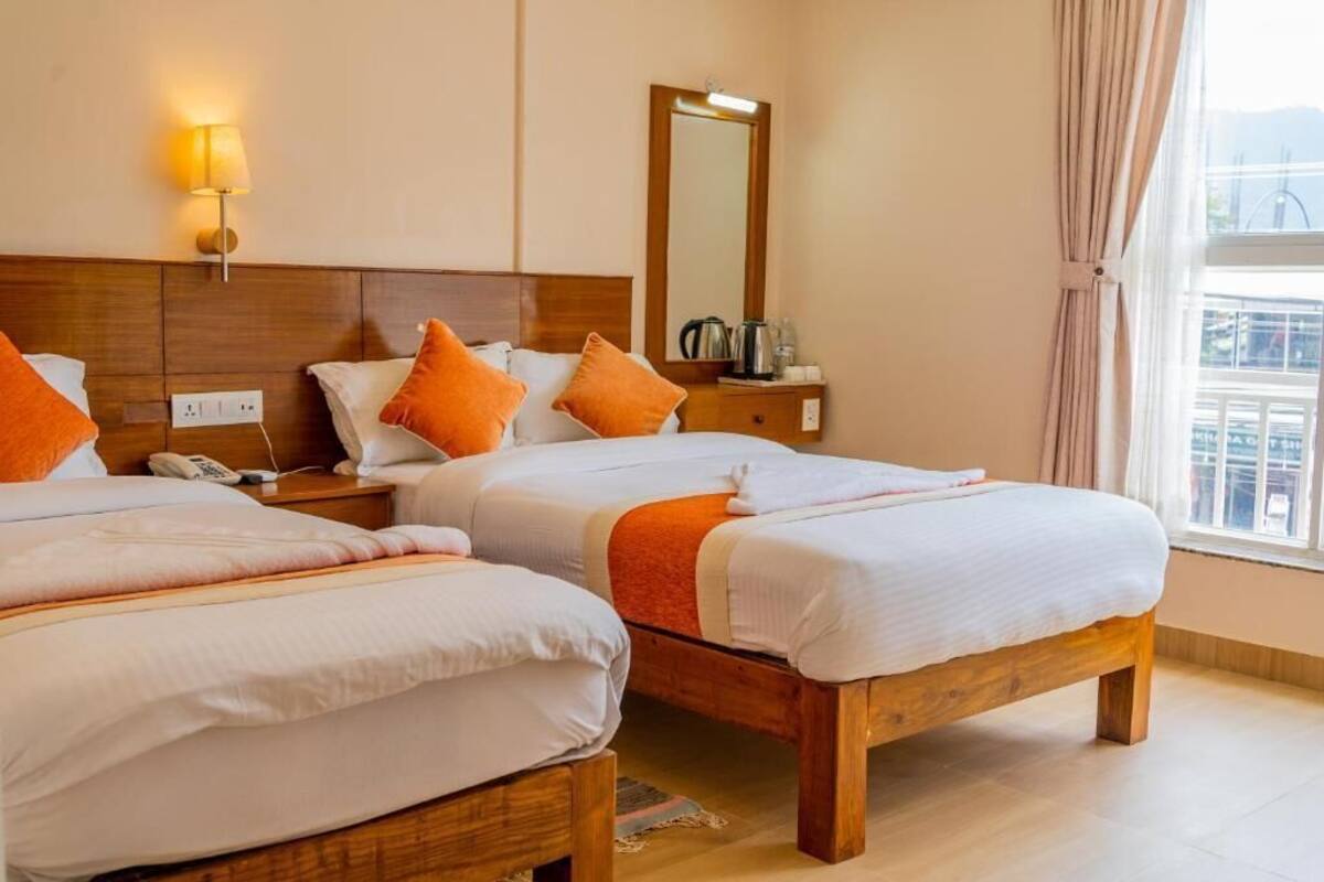A hotel room with two beds, a wooden headboard, a nightstand with a lamp, a mirror, and a window with curtains.