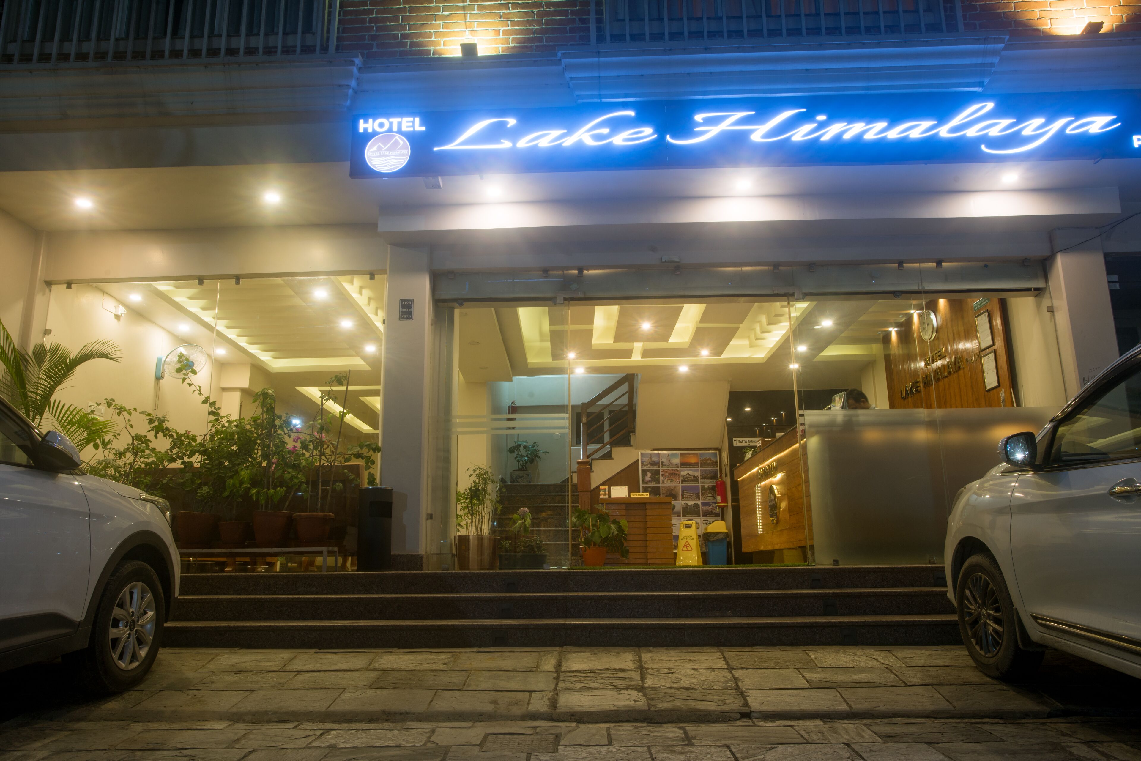 Exterior del Hotel Lake Himalaya, con diseño moderno, que presenta un llamativo letrero de neón, una escalera que conduce a la entrada y plantas en macetas alrededor del área de acceso.