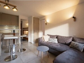 A modern living space with a sofa, a small round table, a kitchen area with a bar stool, and a doorway leading to another room.