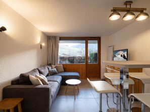 A modern living room with a sofa, a small round table, a bar area with stools, and a large window with a view of a snowy landscape.