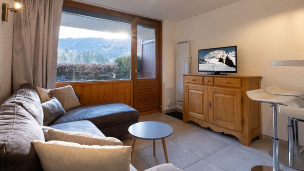 A cozy living room with a sofa, a small round table, a cabinet, and a television on the wall. - Cozy 2-room flat, 4 guests, near slopes, garage (La Clusaz)