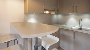 A small kitchenette with a white countertop, a sink, and a stove. There are two white bar stools and a small table. - Cozy 2-room flat, 4 guests, near slopes, garage (La Clusaz)