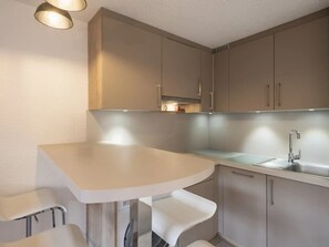 A small kitchenette with a white countertop, a sink, and a stove. There are two white bar stools and a small table. - Cozy 2-room flat, 4 guests, near slopes, garage (La Clusaz)