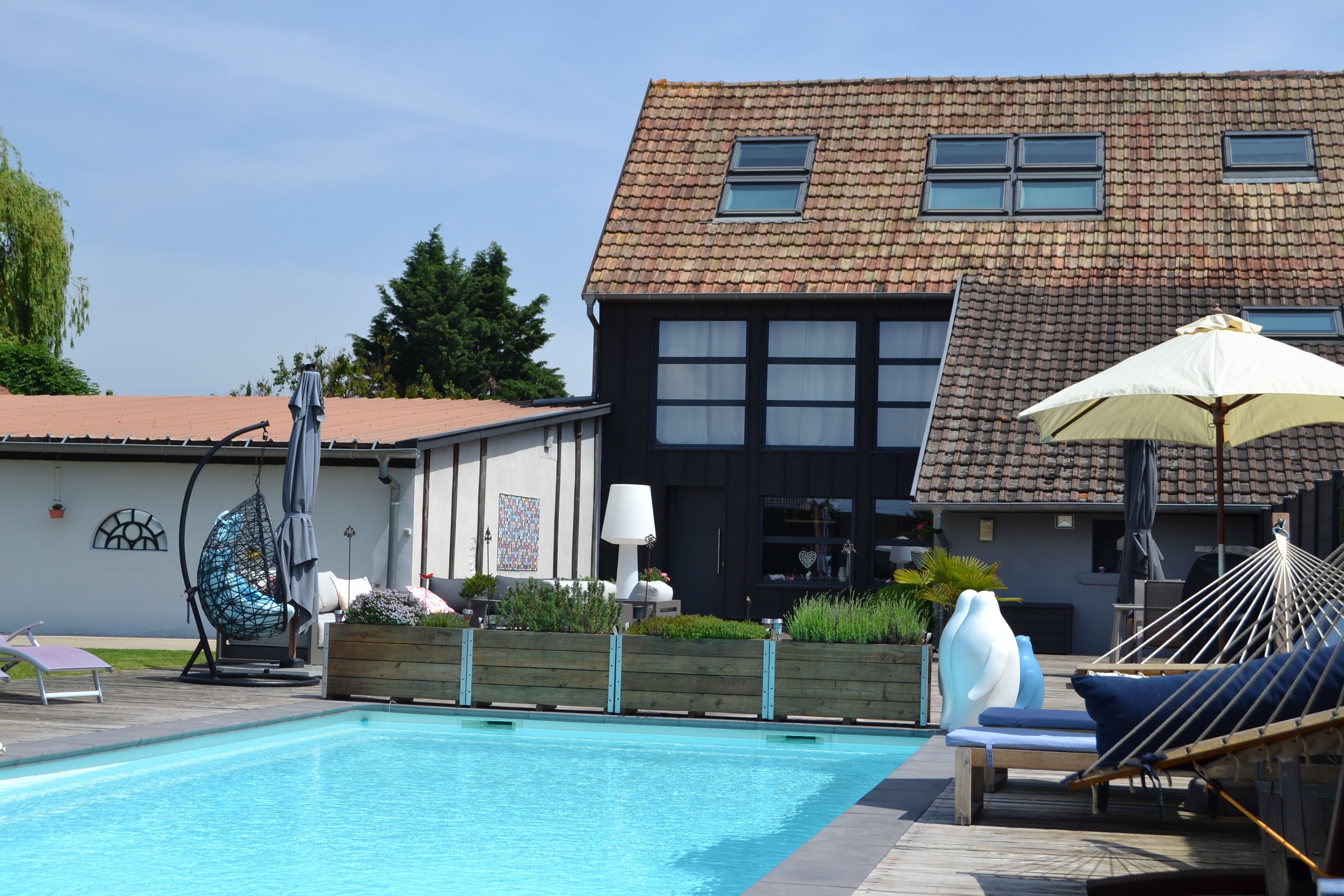 A swimming pool with lounge chairs and umbrellas, a wooden deck, and a two-story building with a tiled roof.