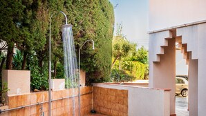 Un área de ducha exterior moderna con suelo de baldosas, barandales de metal, rodeada de follaje y un muro de piedra.