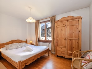 A wooden bed with white linens, a wooden wardrobe, a wicker chair, and a window with curtains. - Charming central apartment with WIFI and parking in Chamonix (Chamonix-Mont-Blanc)