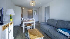 Un salon moderne avec un canapé gris, une table basse en bois, une table à manger avec des chaises, un téléviseur posé sur un meuble et une lampe sur une étagère.