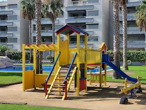 A playground with a slide, climbing structure, and swings, surrounded by palm trees and a building in the background.