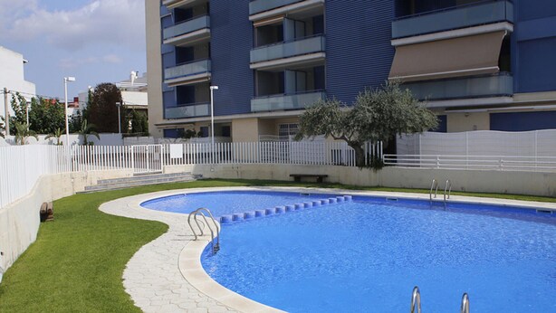 A swimming pool with a curved edge, surrounded by a white fence, and adjacent to a multi-story building with balconies.